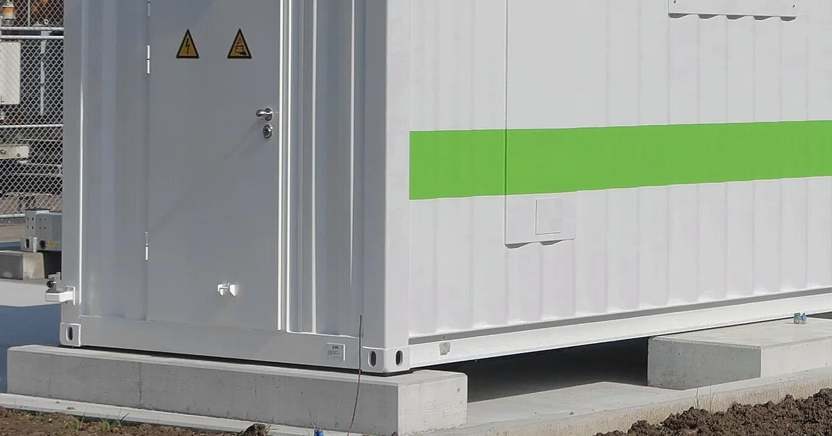 Corner fitting and base rail of a white battery storage container resting on concrete blocks, door with warning signs beside it